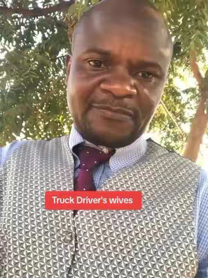 A medium close-up shot of a man with a shaved head and a slight smile. He's wearing a light blue shirt, a patterned grey vest, and a purple patterned tie. A red rectangle at his chest says 'Truck Driver's wives'. The background is a blurry mix of green foliage and tree branches.