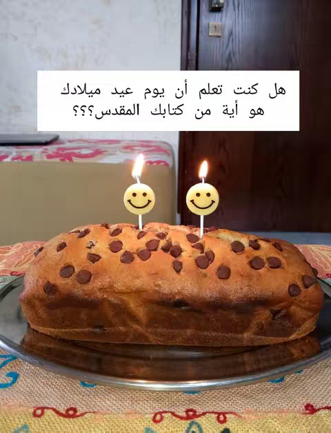 The image shows a cake decorated with chocolate chips and two smiley face candles. Inside of a white rectangle is arabic text saying "Did you know your birthday verse is from the bible? Write your birthday in the comments and I will tell you your special verse. Share the idea with your friends so they can know their verse too"