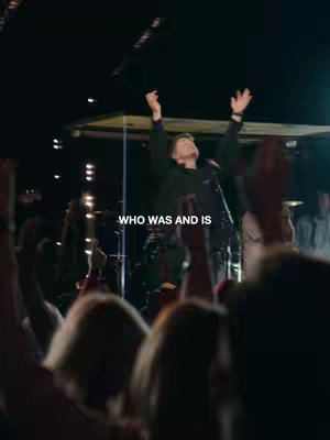 A live performance scene with a man on stage in the center, arms raised in worship. He is wearing a dark top. The stage is lit with spotlights. The audience is visible in the foreground, their arms raised as well. Text overlayed on the image reads “WHO WAS AND IS”.