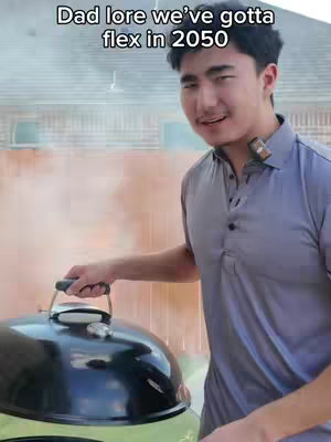 A young man, standing outdoors, smiles towards the camera while holding the lid of a black grill. He wears a gray polo, and in the background is a house, with smoke rising.