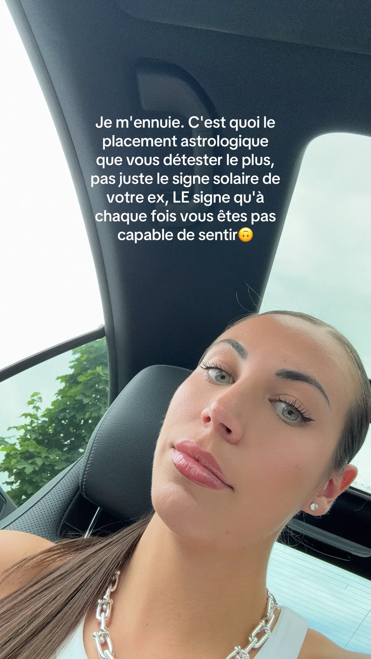 A selfie taken from inside a car, featuring a woman looking at the camera. The woman's face is the primary focus, with a text overlay at the top. She has light skin, long brown hair, and is wearing a white top and a silver chain necklace. The background is blurred, with hints of greenery and the car's interior.