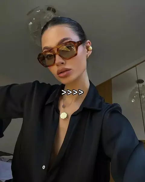 A woman wearing brown sunglasses and a black blouse is positioned centrally in the frame. The woman has light skin, dark hair, and is wearing a gold earring. The image is well-lit, with a neutral background that suggests an indoor setting. The primary color scheme is neutral with the black blouse and brown sunglasses.