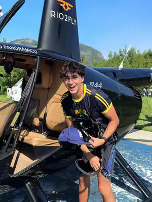 A young man stands in front of a helicopter, smiling at the camera. He wears a navy blue and yellow shirt, black shorts, and holds a baseball cap. The helicopter is black with the words "RIO2FLY" on the side.