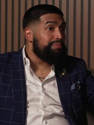 A medium close-up shot focused on a Black man. He is wearing a dark blue blazer over a white dress shirt and has a gold chain around his neck. His hair is neatly styled with a fade. The background is a neutral color, with vertical lines.
