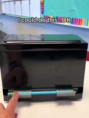 A black rectangular box on a white surface, with a hand pointing at a pencil that's in a lower compartment of the box. The background is blurred, showing a classroom or office setting. Text overlay at the top reads 'I could do this... OR'
