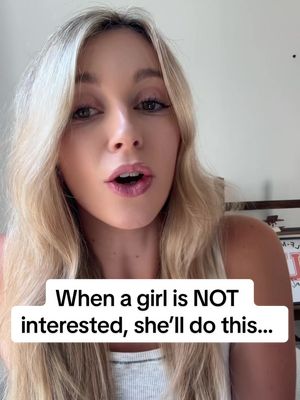 A medium shot of a young, blonde woman with her mouth slightly open, suggesting speech, against a blurred, neutral background. She's wearing a light tank top. A white text box at the bottom reads: "When a girl is NOT interested, she'll do this..."