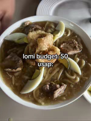 A close-up shot of a bowl of lomi noodles, with pieces of meat, boiled eggs, and crispy fried pork rinds. The bowl is on a surface partly visible at the bottom of the frame, with a white plate in the background.