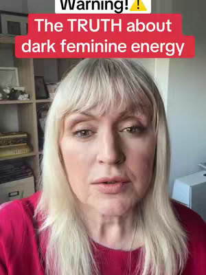 A woman with pale skin and blonde hair looks directly at the camera. She wears a red top and has light makeup. A shelf with various items is visible in the background, along with a white box on the right.
