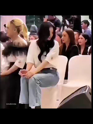 The image captures a scene at an event, likely a public gathering or press conference. Two women with dark hair are the focus, seated and facing forward. The woman on the left has her hair styled and is wearing a dark top with a decorative neckline. Beside her on the right, another woman has black hair, a white top, and jeans, partially turned, seemingly engaged with events. The background comprises additional people, a stage or presentation area, and professional lighting.