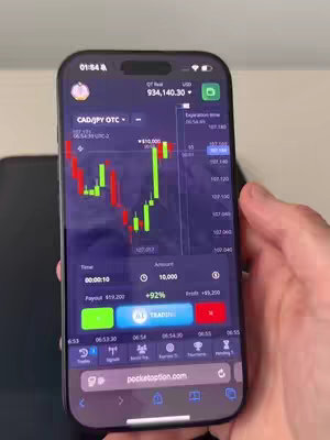 A phone, displaying a trading app, is held by a person with visible hands. The app screen shows a currency chart and trading interface.