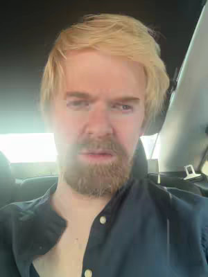 A medium shot shows a man with blonde hair and a beard from the chest up, inside a car. He has a furrowed brow and is looking towards the camera with a slightly distressed expression. The background shows the interior of the vehicle, including the headrest of the seat and part of the window. The man is wearing a dark shirt with buttons.