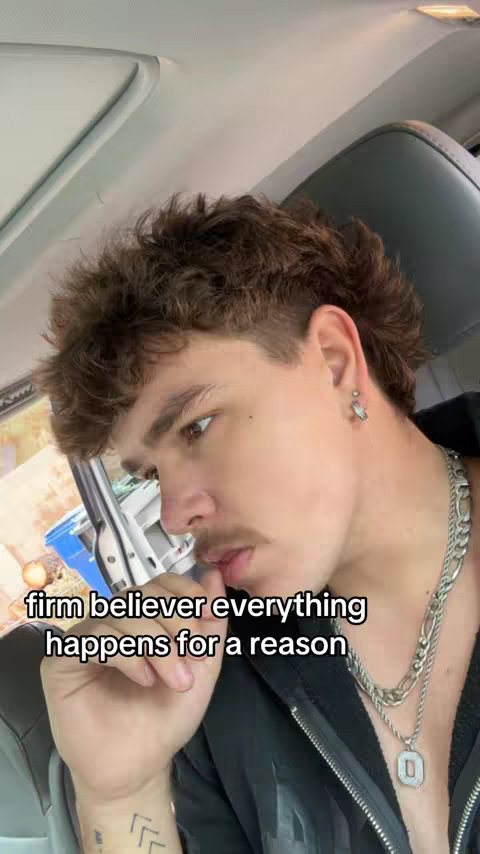 A young man with a mullet haircut and a small mustache is seated inside of a car. The background shows the interior of the vehicle, including the seat and windshield. The man is looking away from the camera, with text overlaid on the image, indicating a message about believing that everything happens for a reason.