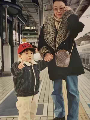 A full-length, slightly aged photograph taken at a train station. A young child wearing a red baseball cap and denim jacket is facing the camera with a pointing gesture. An older woman with glasses and a black coat with leopard-print trim is holding the child's hand. They are standing on a platform, with a train visible in the background.