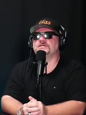 A man with a beard, wearing sunglasses, a black t-shirt, and a cap that has GG33 written on it, sits behind a microphone while wearing headphones.