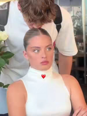 A woman with light skin is in the foreground, looking slightly to the side with an ambiguous expression. She wears a white, high-necked top. Behind her, a man is leaning over her, his face obscured, seemingly about to kiss her head. The color palette is natural, with soft focus in the background and a slightly out-of-focus floral arrangement on the left.