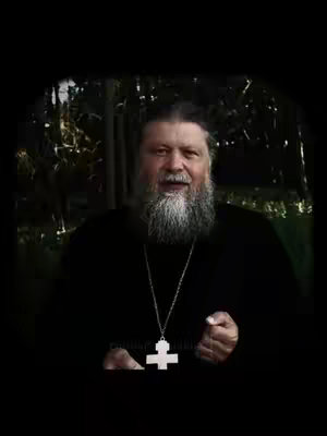 A man with a long gray beard is centered in the frame, wearing a black garment and a silver chain with a large white cross pendant. Dark, blurred foliage forms the backdrop, and the overall lighting is dim, casting shadows.