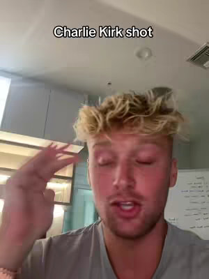 The image is a medium shot of a young, fair-skinned man with styled, blonde hair, positioned with the upper body visible. The man is looking slightly to the left with his eyes closed. His right hand is raised near his head with his fingers spread. The background is a bit blurred and shows elements of a bright indoor setting with a ceiling and a visible vent. Above the man, a text overlay reads 'Charlie Kirk shot' in a black, sans-serif font.