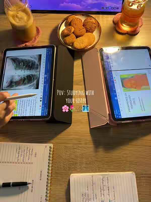 A top-down shot of a wooden table with two open tablets showcasing study materials, accompanied by cookies and drinks, and handwritten notes. The lighting is natural, with a focus on the items. The background features a monitor.