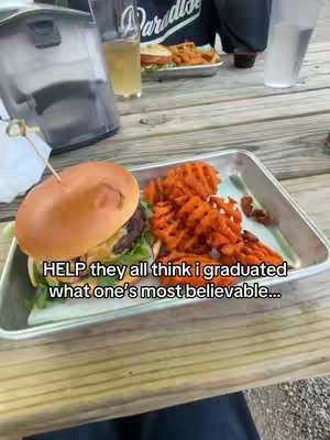 The cover image shows a stainless steel food tray on a wooden table. The tray holds a burger impaled with a toothpick, alongside sweet potato waffle fries. In the background, there are glasses and more food, hinting at a dining setting. The text overlay reads, 'HELP they all think i graduated what one's most believable...'.