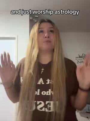 A young woman with long blonde hair stands facing the camera, raising her hands. She's wearing a brown top featuring white text and a cross necklace. Behind her is a white wall with a blurred message and a door frame visible. On the top of the cover text appears the phrase \"and just worship astrology\". 