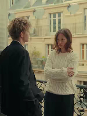 A medium shot of a balcony scene features two people standing close to each other. A woman in a white woven sweater and black skirt is on the right with her arms crossed, looking at the other person. On the left, a man in a black jacket appears to be facing the woman while standing near the edge. Behind them, the balcony's wrought iron rail and a building with many windows are visible. The image is brightly lit, with soft natural light creating a warm tone.