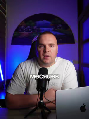 A man with short hair, a white t-shirt, and a microphone sits in front of a laptop. The background features a blurry, colorful abstract design and blue accent lighting. Text in Russian is added to the image.