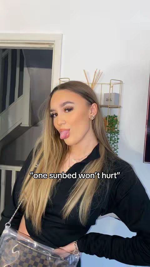 A woman with long blonde hair and full makeup is sticking her tongue out. She's wearing a black top, and there's text overlay on the image reading, "one sunbed won't hurt". The background is a home interior with a staircase on the left side.