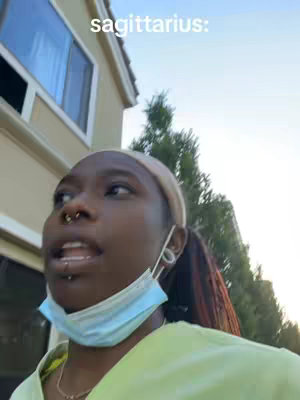 A woman with dark skin and a nose piercing stands outside, partially obscured by a pale yellow hoodie and sky blue surgical mask. Text across the top of the frame reads "Sagittarius:" She's looking slightly off-camera with a focused expression.