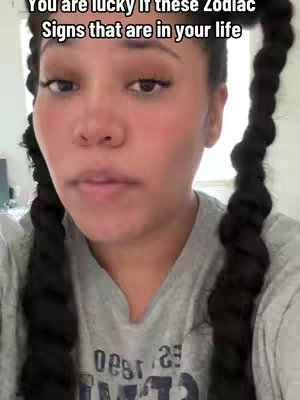 A selfie-style video with the creator's face filling the frame. She has dark hair styled in two long braids. The background is a blurry indication of a room, likely indoors. The lighting appears to be natural, and the creator is looking at the camera with a neutral expression. The color palette is muted, with the focus on the creator's skin tone and clothing.