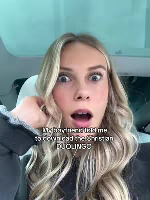 A woman with blonde hair looks directly at the camera with wide eyes and an open mouth, a surprised expression. Text overlays at the bottom state, 'My boyfriend told me to download the Christian DUOLINGO'. The background suggests a car interior. The overall composition is a selfie, framed to capture the woman's face and upper body, with a focus on her expression.
