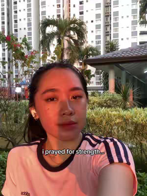 A young woman with dark hair and fair skin is centered in a selfie-style shot. She's wearing a white t-shirt with blue and black stripes on the sleeves. The text overlay reads 'i prayed for strength...'. The background includes a blurred cityscape with tall buildings and greenery.