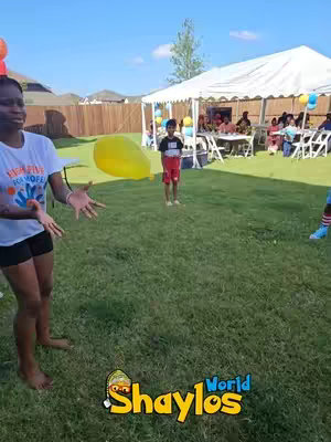 A woman with short hair stands in a grassy backyard with arms outstretched, near a large yellow balloon. A child in a red and black shirt stands further back, with a white tent and a group of people visible in the background. The sky is blue. The bottom corner of the image includes a yellow logo that says Shaylos World. 