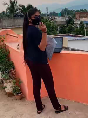 A woman stands on a rooftop, wearing a black mask and casual clothing, with a laptop open nearby and a city backdrop.