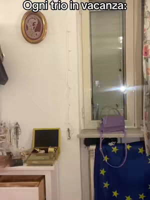 An interior shot featuring a white wall with a framed oval picture, a window, and a blue skirt with yellow stars. On a white surface, there is an open box with various trinkets.