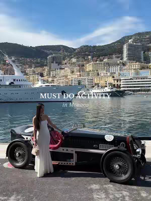 A woman in a white dress stands beside a vintage car in Monaco, with a harbor and city buildings in the background. The sky is blue with some clouds.
