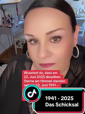 A woman with light skin and dark hair gazes directly at the camera. Text overlays appear in red boxes: one with statements about the alignment of stars and another stating '1941-2025 Das Schicksal'. The backdrop is a dimly lit room with indistinct objects and makeup. The shot has focus on the woman's face.