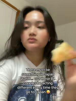 A woman with dark hair is shown in a selfie-style video. She is wearing a white t-shirt. Text overlaid on the image reads, 'There must be something in Japan water... cuz these BL couples be acting different every time they go to Japan'. She is holding a piece of food with a blurry background.