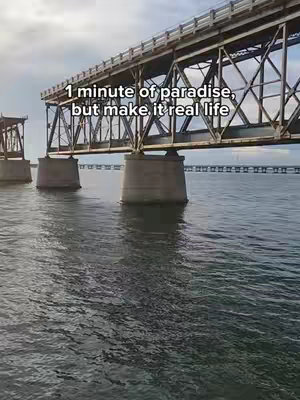 A wide shot of a bridge over water under a cloudy sky. The bridge is made of metal and concrete, and its supports stand in the water. The water is choppy, and the sky is filled with clouds.