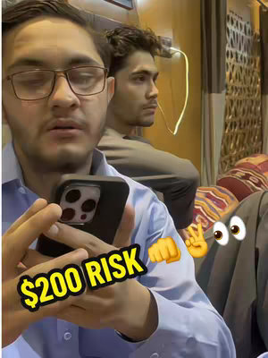 A selfie shot with two young men. The main subject, wearing glasses, is holding a phone displaying a black screen with a camera island. The text overlay says "$200 RISK". An animated cartoon eye and fist emoji add to the playful theme. The background includes a glimpse of a fancy interior or restaurant setting.