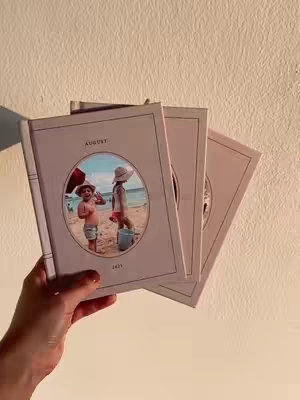 A hand holds three photo books against a pale, textured wall. The books are a soft, muted pink with a minimal design. Each book features an oval photo on the cover showcasing children on a beach with text above and below the picture. The books are stacked, with one in focus and the other two slightly blurred.