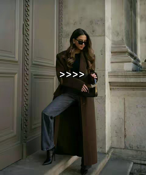 A woman wearing sunglasses, a long brown coat, and jeans stands on steps in front of a building. The image has a neutral color palette with shades of brown. There are text overlays with arrows >>>>>.