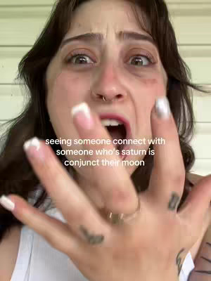 A close-up shot of a young woman with dark hair and a piercing. She is looking at the camera with wide eyes and an expression of shock or distress. Her hand is raised and closer to the camera. Her lips are parted slightly. The background is a blurred light green.