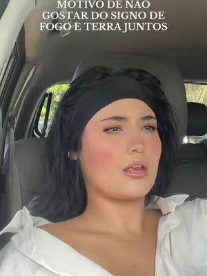 A woman is seated inside a car, angled towards the camera. She is wearing a white shirt and a black headband. Above her, text in white is partially visible. The background includes the car's interior and sunlight.