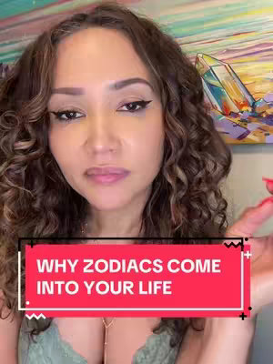 A woman with long, curly brown hair looks directly at the viewer. She has makeup on, including eyeliner and lipstick. A red rectangular text box at the bottom reads, "WHY ZODIAC COME INTO YOUR LIFE." A vibrant painting and crystals are in background.