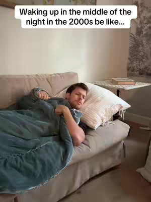 A man is lying in a bed covered with a blanket. The man is facing the camera. The man is wearing a grey t-shirt. There is a white pillow behind him. There is a small table in the background with a book on it.