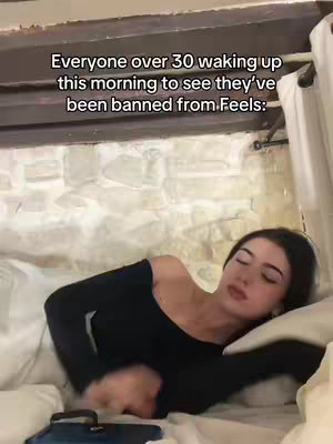 A woman lies in bed, eyes closed, appearing to be asleep or waking up. The room is softly lit with muted tones; a headboard and curtains provide the visual background.
