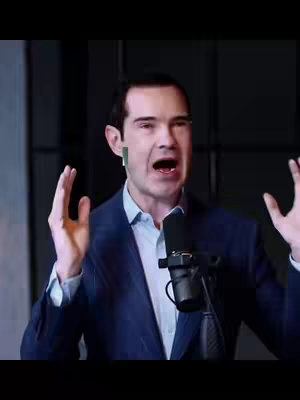 A man in a dark suit and light blue shirt is speaking into a microphone, with his arms raised open. The background is a blurred, dark setting.