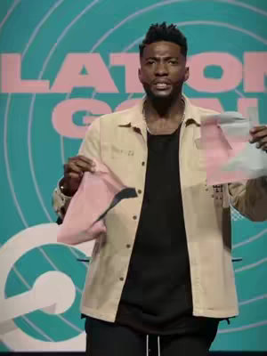 A man with short black hair stands in front of a teal background with pink text. He is wearing a dark t-shirt under a tan button-down jacket and black pants. He is holding up two pieces of pink paper in his hands, looking directly at the camera.