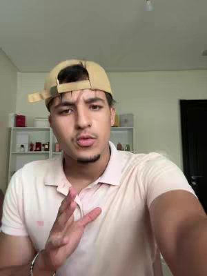 A man wearing a beige cap and a light pink polo shirt is gesturing with his right hand in front of a neutral background. The lighting is soft and natural.