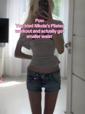 A woman in a mirror selfie with a caption overlay. She's wearing a white shirt and denim shorts, posing from the waist up. A text overlay reads "Pov: You tried Nikola's Pilates workout and actually got a smaller waist."